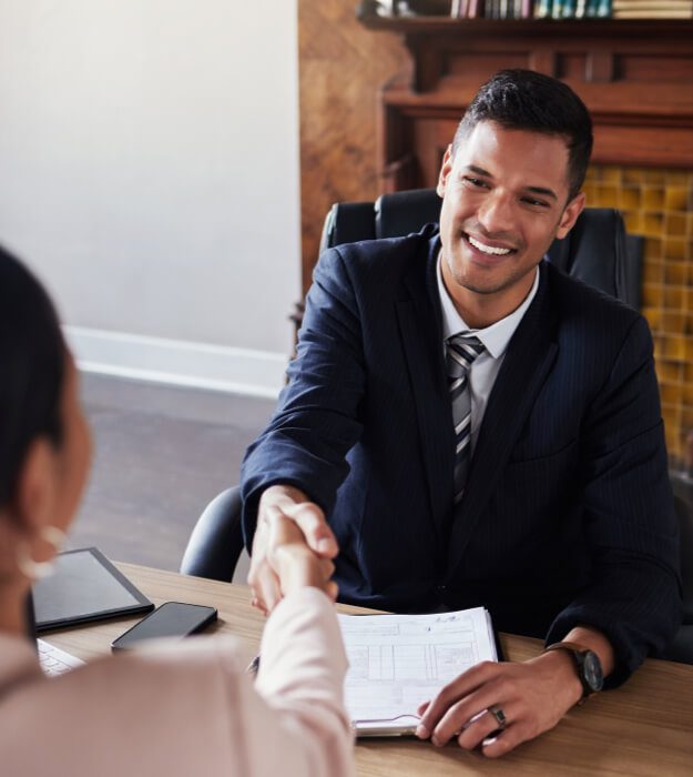 A man in a suit shaking hands with a woman across his desk with paperwork in front of him