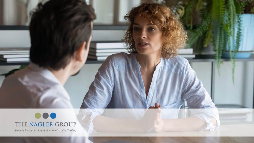 a woman with curly red hair speaks to a colleague across the table from her in an office setting